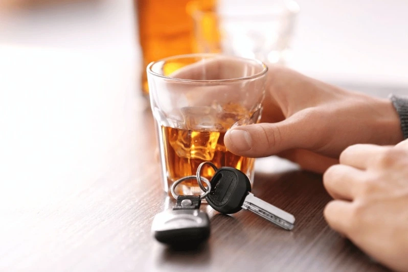 Close-up of a person holding a glass of amber-colored alcoholic drink (whiskey or liquor) with a set of car keys resting on the table directly in front of the glass, symbolizing drinking and driving.