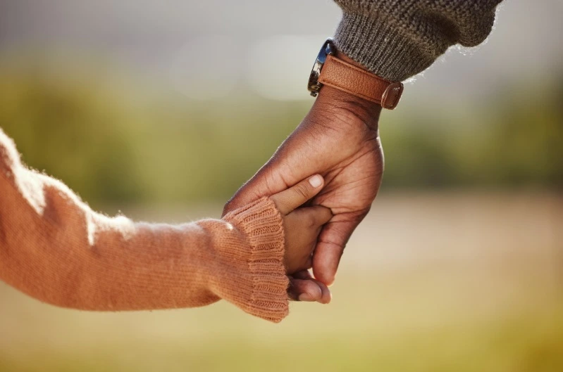 Close-up of a dark-skinned adult's hand, wearing a watch with a brown leather strap, gently grasping the tiny hand of a child wearing an orange knit sleeve, suggesting guidance or care.