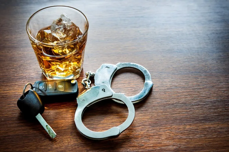 A glass of liquor with ice, a set of silver metal handcuffs, and a car key fob resting on a dark wooden table, symbolizing a DUI arrest or charge.