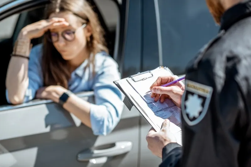 A female driver looking distressed and holding her head while a police officer writes a traffic ticket or citation on a clipboard during a traffic stop.