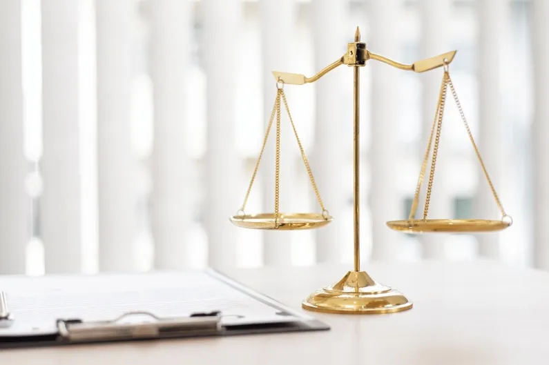 A brass or golden scale of justice standing next to a clipboard with legal papers on a white desk, symbolizing law and fairness.