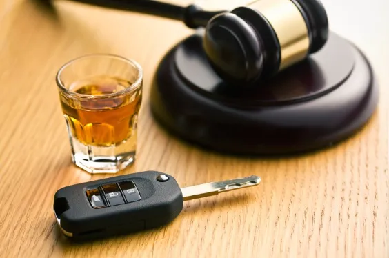A shot glass of liquor and a car key fob next to a judge's gavel on a wooden desk, symbolizing the legal consequences of drinking and driving (DUI).