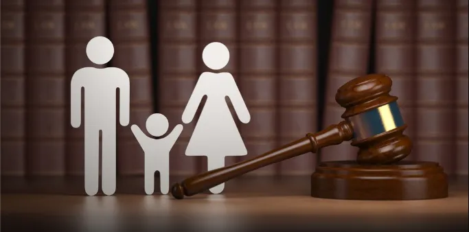 A wooden judge's gavel resting next to white stick figures representing a man, a woman, and a child, set against a background of leather-bound "LAW" books. This image symbolizes family law, custody, divorce, or legal proceedings involving families.