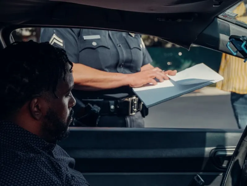 A close-up, side-view image of a serious-looking man (driver) seated in a car while a police officer, wearing a uniform with sergeant stripes, stands outside the car holding a clipboard or ticket book. The image depicts a routine traffic stop or the issuan