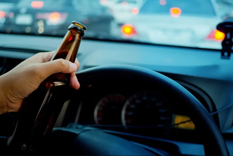 A close-up image from inside a car showing a person's hand holding an open, brown glass beer bottle near the steering wheel and dashboard, with the brake lights of other cars visible through the windshield in traffic. This image symbolizes driving under th
