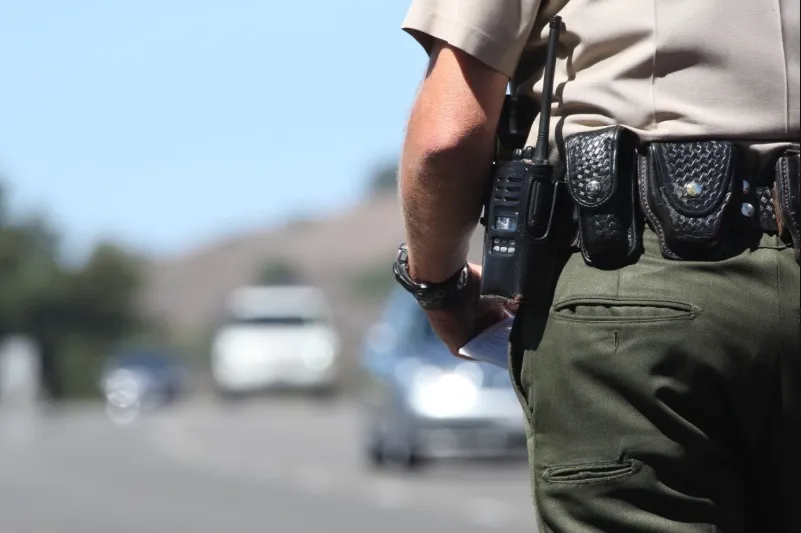 A close-up, back view of a police or sheriff's officer in a tan uniform and dark green pants, standing on the roadside. The focus is on the officer's utility belt, which holds a radio and pouches, with blurred vehicles visible in the background, suggesting