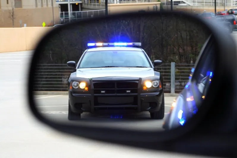 The front view of a black police car with blue emergency lights flashing on the roof, visible in the rearview or side mirror of another vehicle. This symbolizes a traffic stop, a pursuit, or being pulled over by law enforcement.