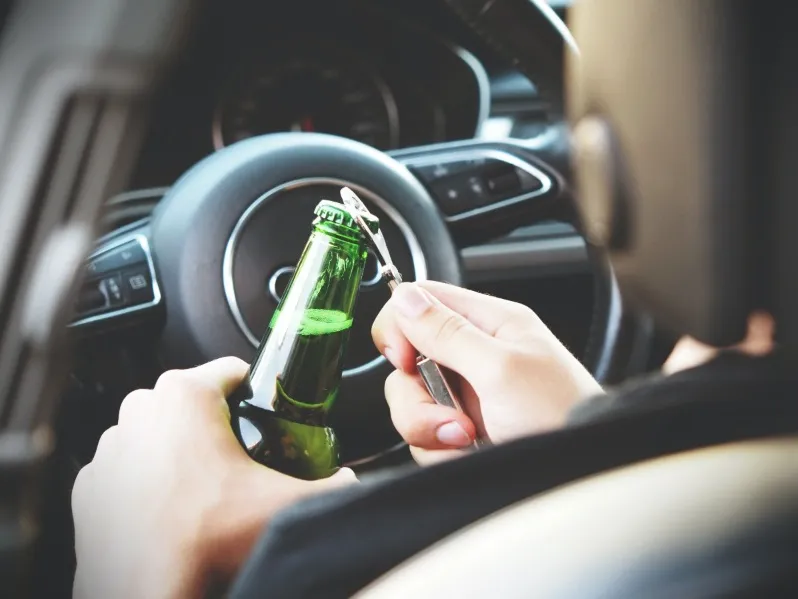 A close-up, over-the-shoulder shot of a person driving a car, using a bottle opener to open a green glass beer bottle near the steering wheel. This image illustrates the dangers of driving under the influence (DUI) and open container laws.