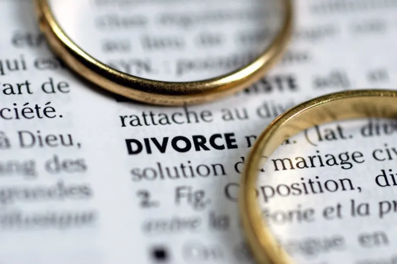 A macro close-up image showing two gold wedding bands resting on an open French dictionary page, highlighting the bolded word "DIVORCE" and its definition. The image visually represents the end of a marriage or the legal process of divorce.