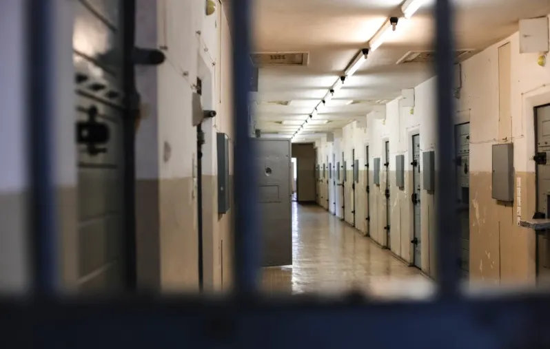 A long, dimly lit institutional hallway of a prison or jail cell block, viewed through steel bars in the foreground. Cell doors line the corridor, emphasizing confinement, incarceration, and the consequences of criminal conviction.