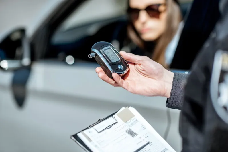 A close-up shot of a police officer's hand holding a digital breathalyzer device with a BAC (Blood Alcohol Content) reading visible on the screen, while a female driver in sunglasses is seen blurred in the background inside the vehicle. This image represen
