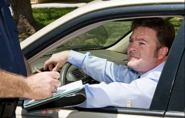 A man in a light blue shirt sitting in his car looks concerned as a police officer's arm reaches in to write a traffic ticket on a clipboard during a traffic stop.
