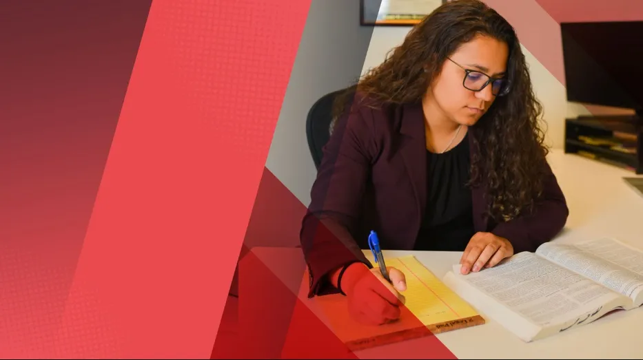 Sabreena Barboza, a student or lawyer with long curly hair and glasses, sits at a desk writing notes on a yellow legal pad next to an open textbook. The image has a decorative red and gray overlay.