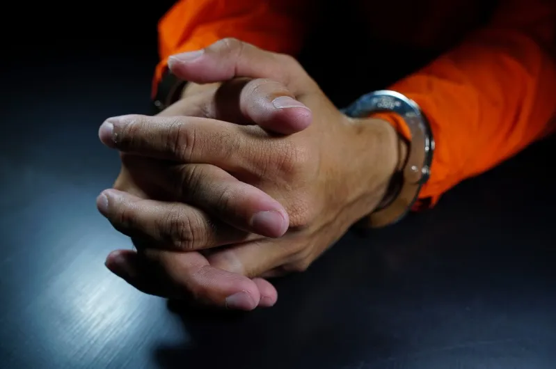 A close-up shot of a person's hands clasped together on a dark table, with a metal handcuff visible on one wrist. The person is wearing an orange inmate or prison uniform.
