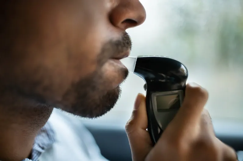 Close-up of a bearded man blowing into the mouthpiece of a handheld breathalyzer (alcohol testing device) indoors or in a car.
