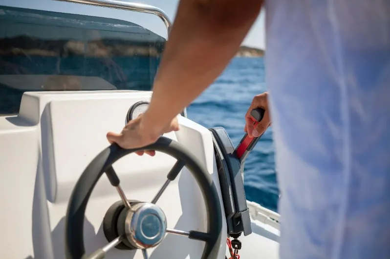 Close-up of a person's hands steering a white speedboat with a black wheel and operating a red-handled throttle lever on a sunny day with blue ocean water in the background.