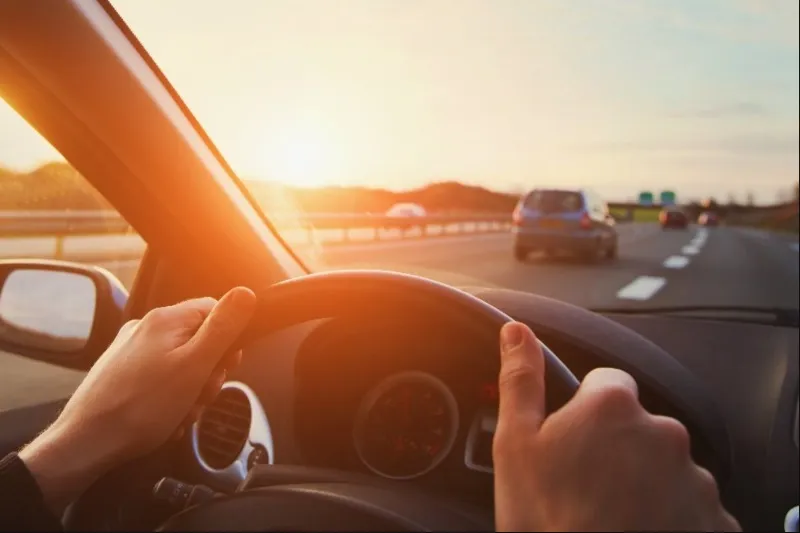Close-up of a driver's hands on a steering wheel, looking out onto a highway with the sun setting brightly on the horizon, creating a lens flare effect.