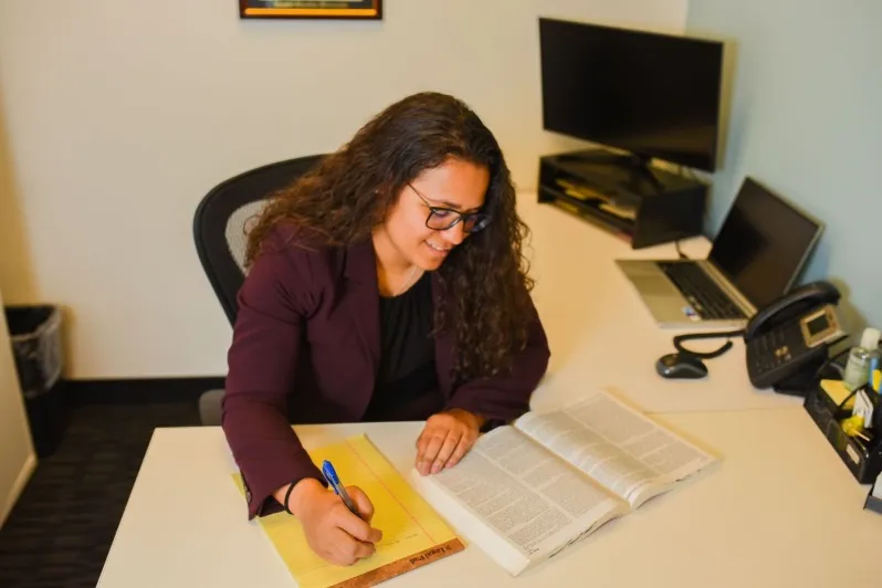 Sabreen Barboza, a young woman with long curly hair and glasses, sitting at a white office desk, writing on a yellow legal pad while looking at an open textbook or law book.