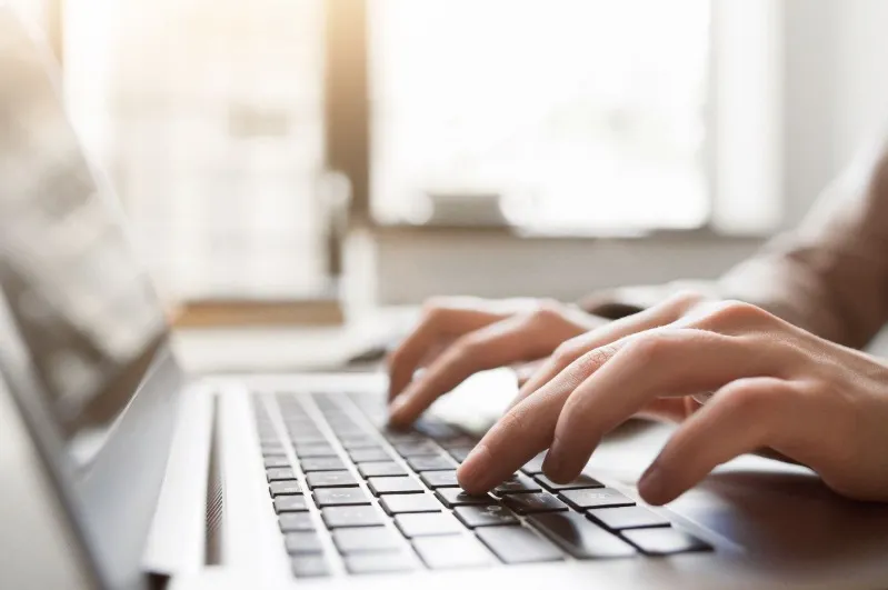 Close-up shot of hands typing quickly on a silver laptop keyboard, with bright sunlight streaming in from a window in the background.