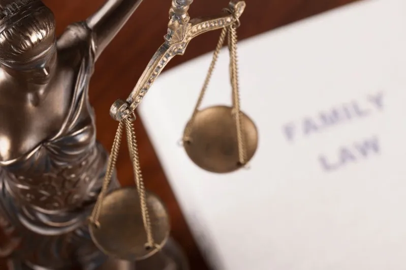 Close-up of the scales of a Lady Justice statue partially obscuring a document that reads 'FAMILY LAW' on a wooden surface.