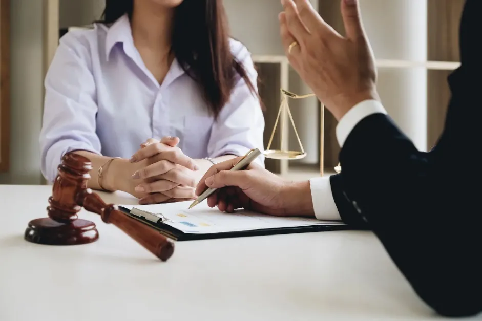 Close-up of a lawyer pointing to a document on a desk with a pen, advising a client. A gavel and scales of justice are visible in the foreground and background, respectively.