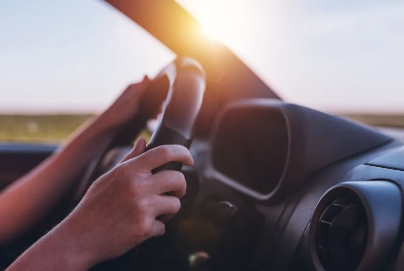 A close-up view from inside a car showing two hands gripping a black steering wheel, driving into a bright sun setting over a clear horizon.