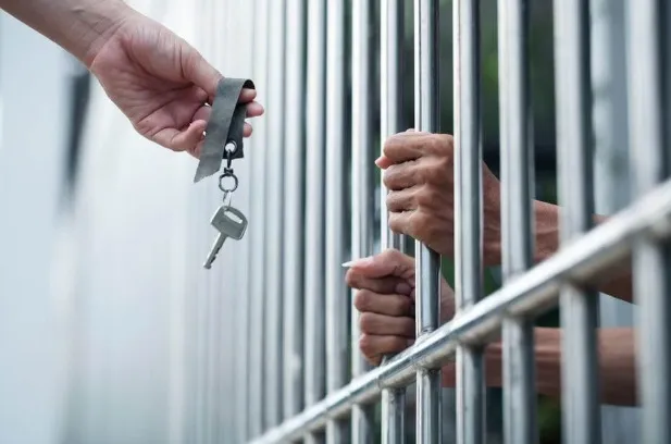 A hand from outside passes a single metal key on a strap through stainless steel prison or jail bars to the hands of an incarcerated person gripping the bars.