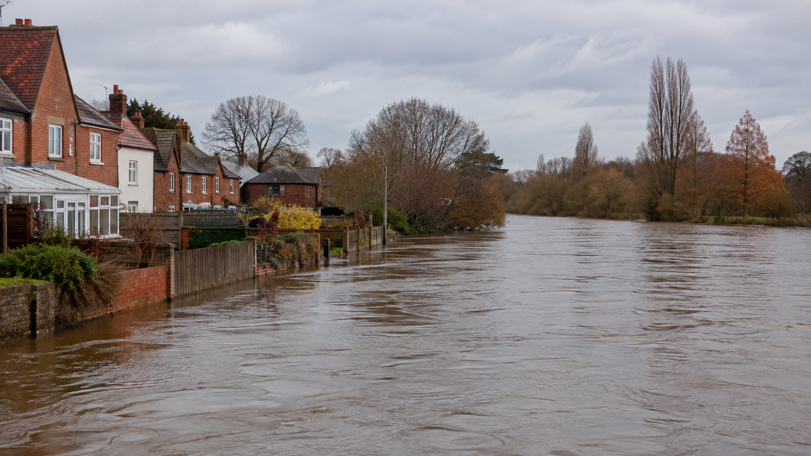 Flooded UK river running high against residential garden fences, showing flood risk affecting nearby houses