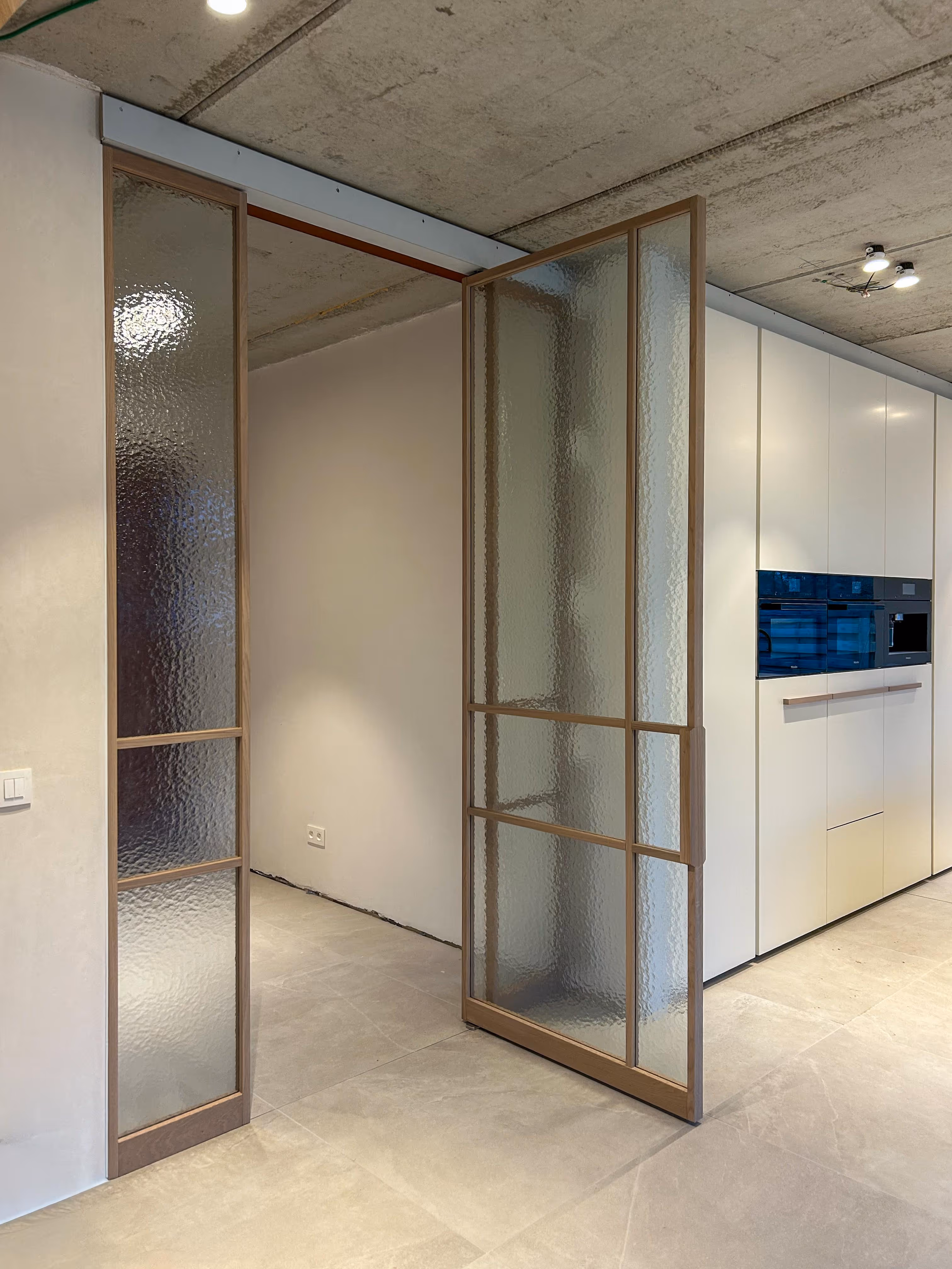 Interieur met een geopende glazen pivotdeur met houten kader, die uitkomt op een kamer met witte keukenkasten.