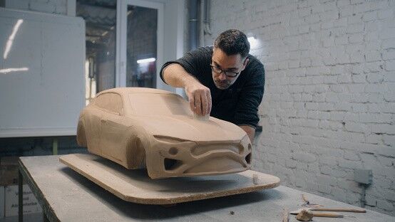 Senior automotive designer sculpts the prototype model of the car made of clay using sculpting tools. Experienced designer works in a workshop. Automotie industry.