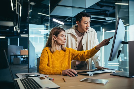 Two freelancers, an Asian man and a woman, work in a modern office, discussing and discussing a joint project, looking at a computer monitor