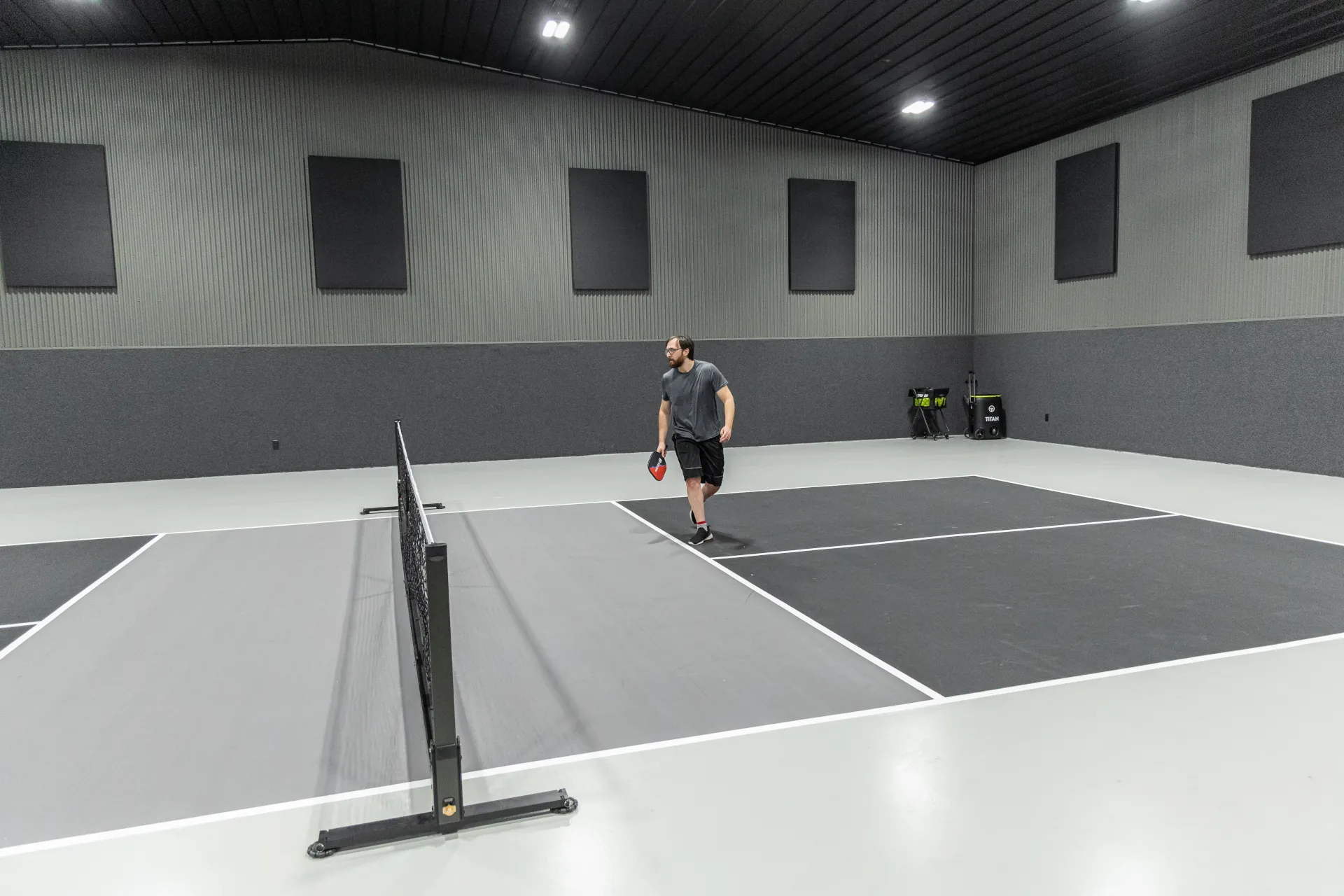 wide view of a man playing on the right side of a black and grey pickleball court