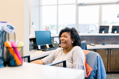 Woman sitting at an office desk laughing, with computer monitors and office supplies in the background.