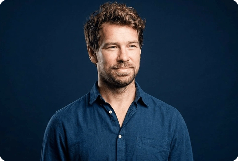 Man with light brown hair and beard wearing a blue button-up shirt against a dark blue background.