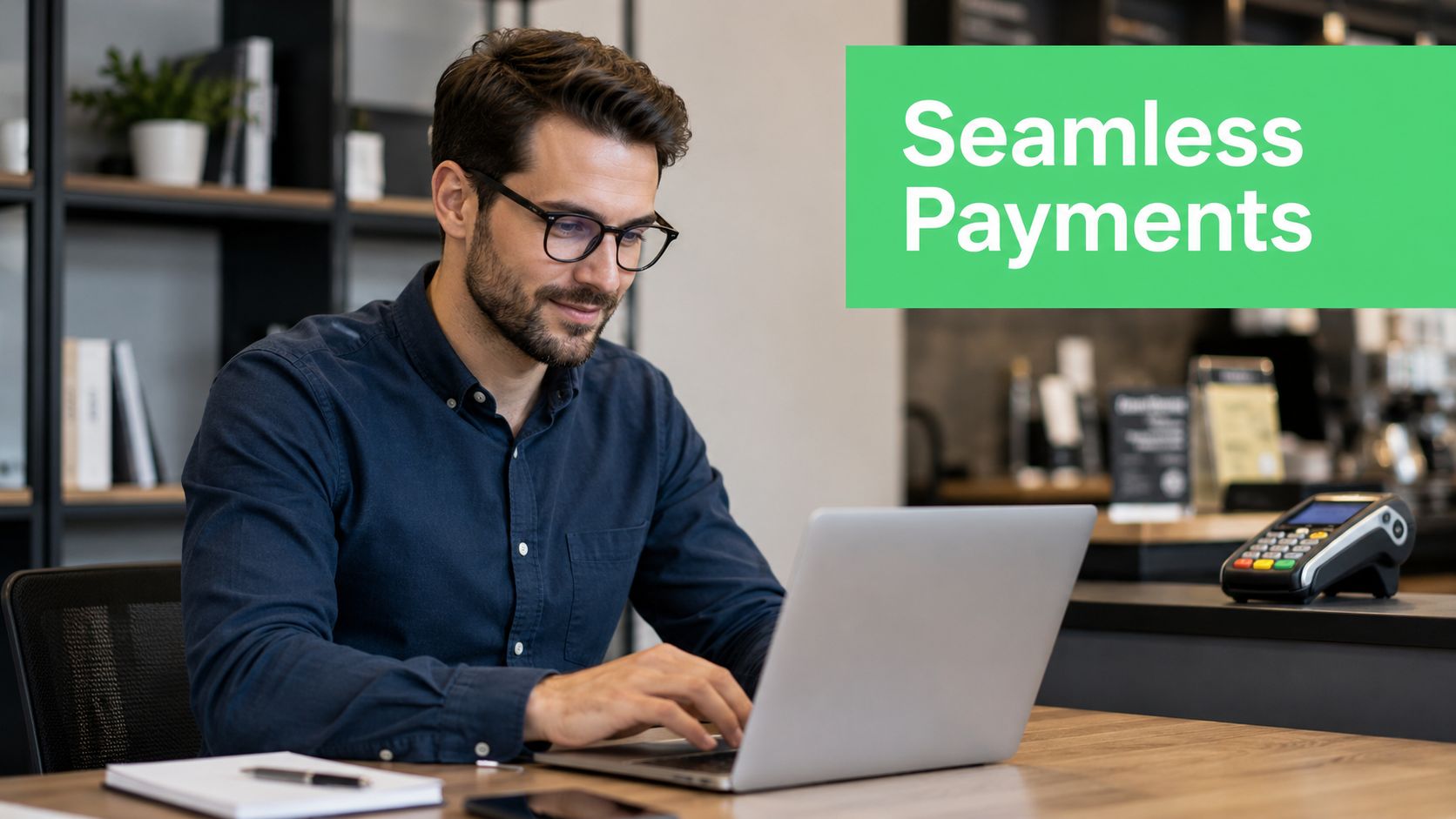 A man wearing glasses working on a laptop at a cafe counter with a payment terminal.