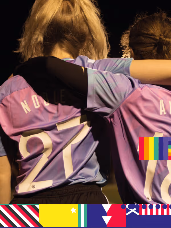 Two players in purple jerseys, 'NOBLE 21' and part of another name visible, hugging on a pitch with a colourful geometric pattern border.