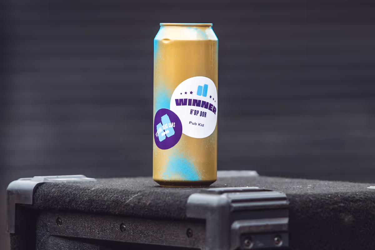 Golden beverage can with 'WINNER H'OP D'OR Pub Kid' and 'FESTIVAL DEUX CANS' stickers, resting on a dark surface with a blurred background.