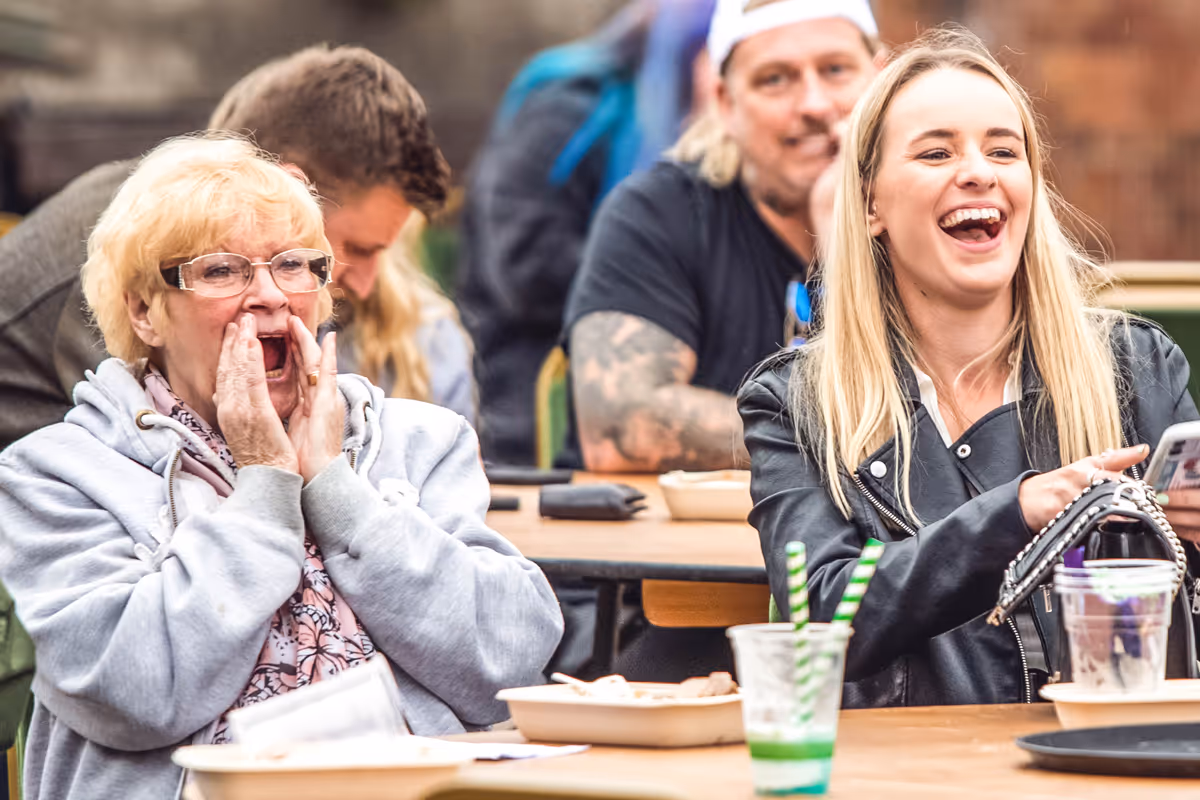 Two women laughing at a table, one older with glasses and one younger looking at a phone, with other people and food in the background.