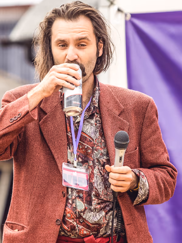 Man with a microphone drinking water, wearing a patterned shirt and blazer, with a purple event background.