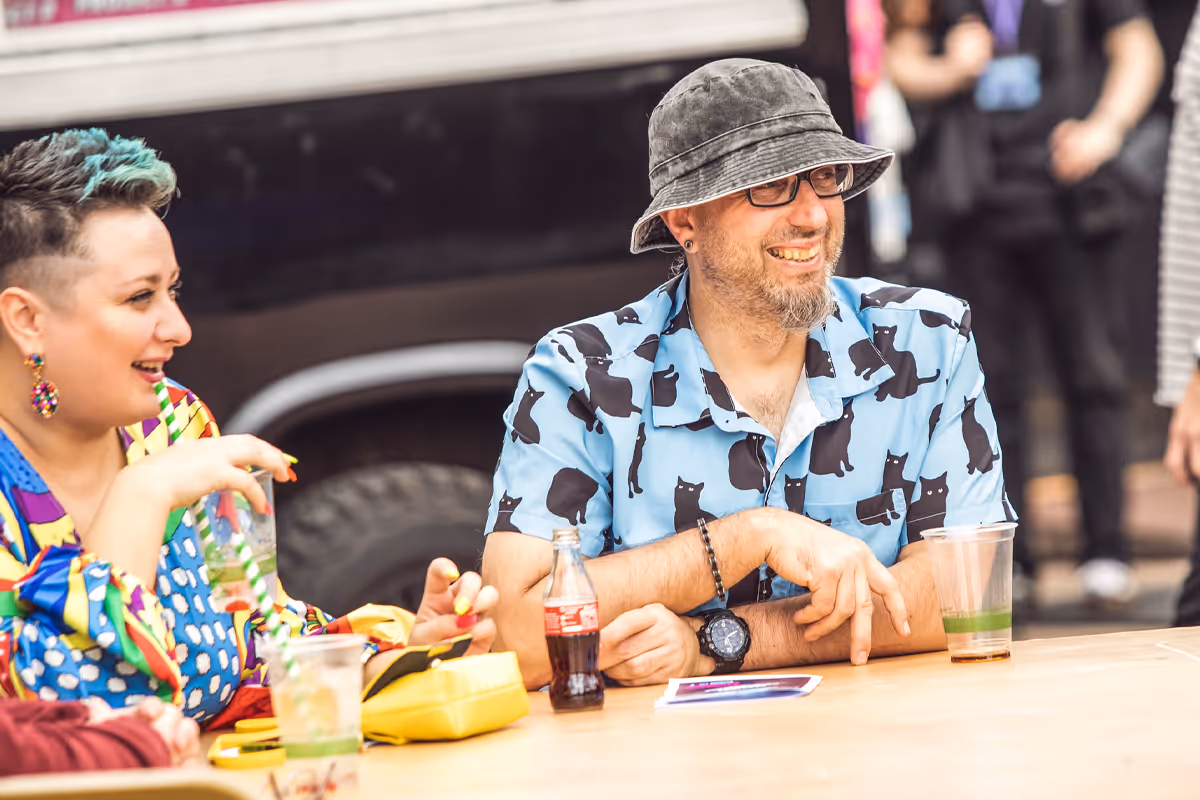 Two people seated at a table laughing, one wearing a colorful outfit and the other in a cat-print shirt and bucket hat, with drinks on the table.