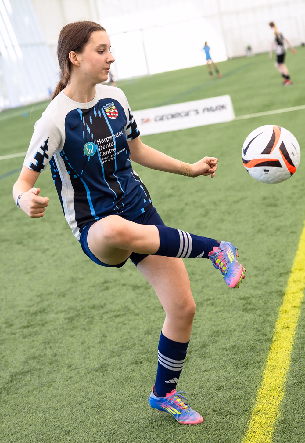 Young female soccer player in blue and white uniform kicking a soccer ball indoors on turf.