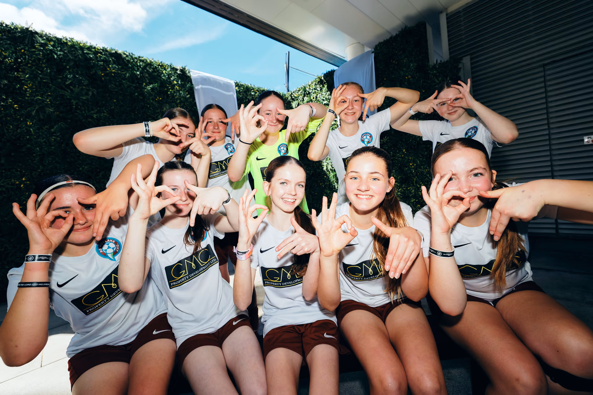 Group of smiling young female soccer players in team uniforms posing with hand signs outdoors.