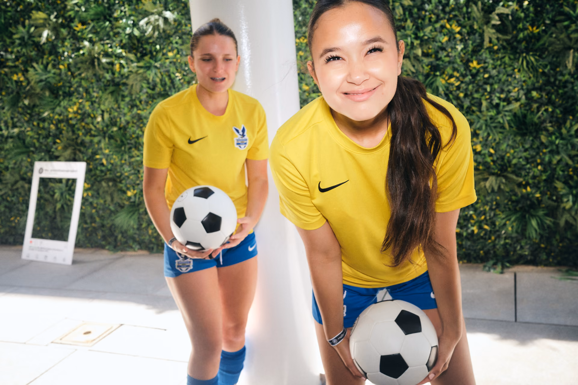 Two young female soccer players in yellow shirts and blue shorts holding soccer balls and smiling.