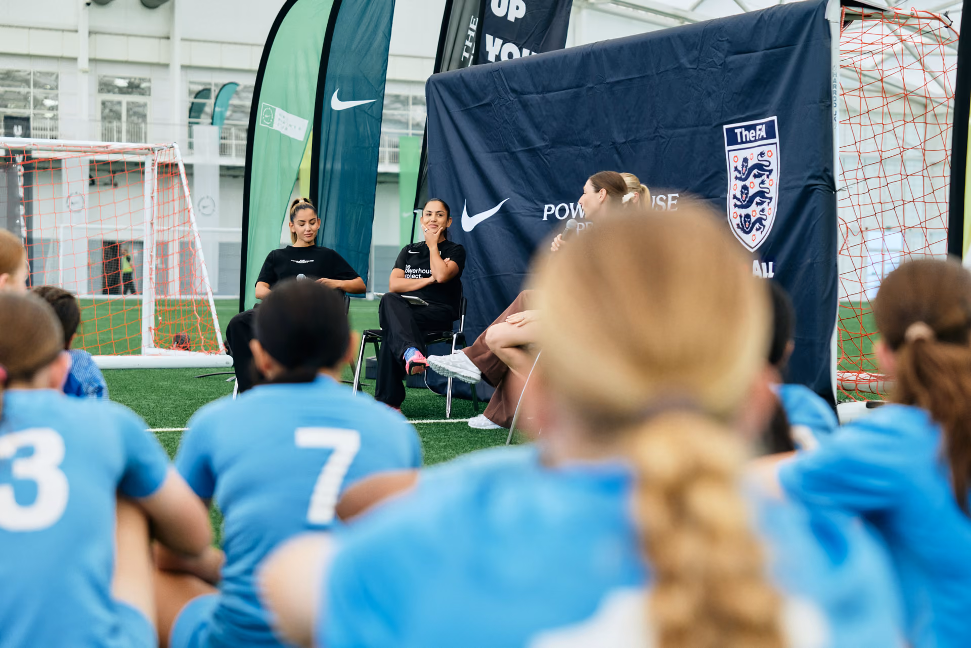 Young soccer players in blue jerseys sit on grass listening to women speakers in black shirts at a sports event.