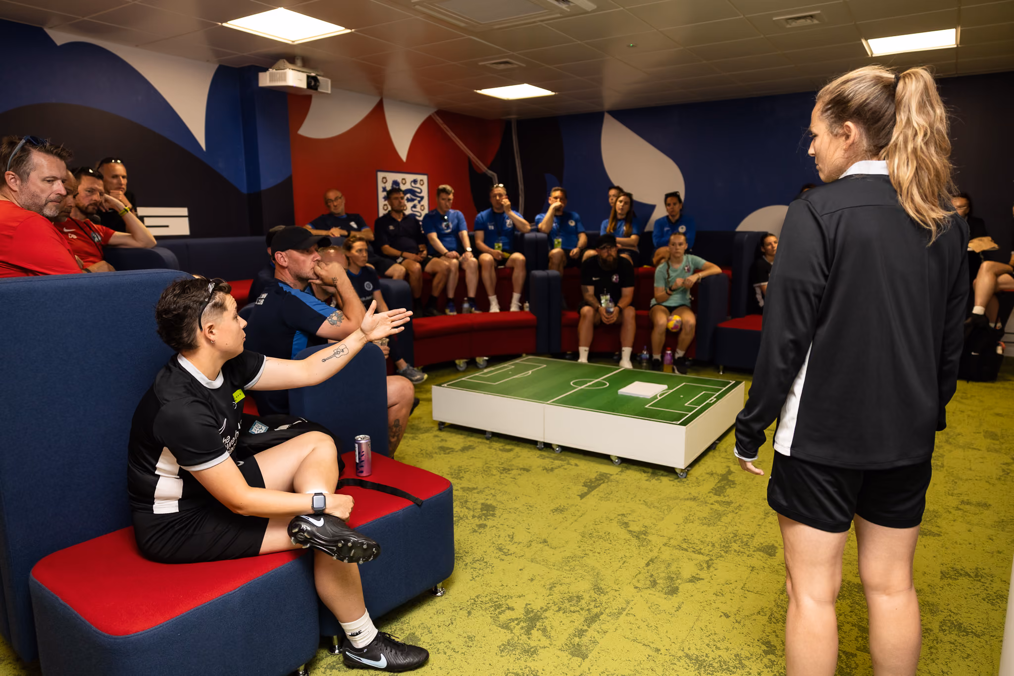 A group of coaches and staff in a meeting room with a soccer tactics board in the center.