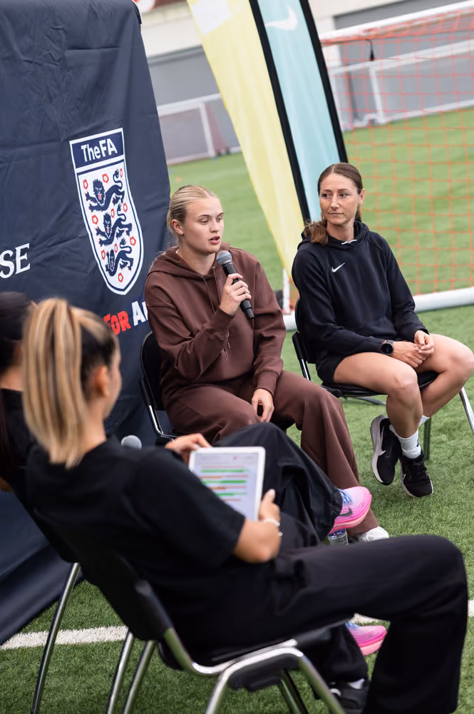 Two women seated on a soccer field with one holding a microphone speaking, another listening attentively.
