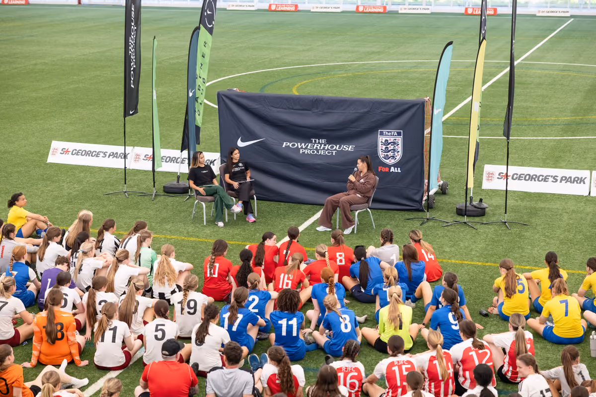 A large group of girls in colorful sports uniforms seated listening to a panel of three on a soccer field.