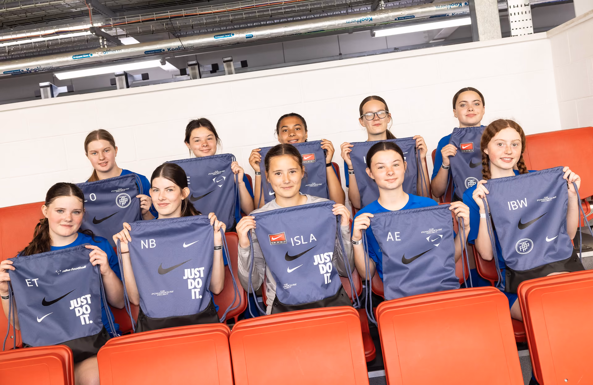 Group of 10 girls sitting in red seats holding navy Nike drawstring bags with initials and logos.
