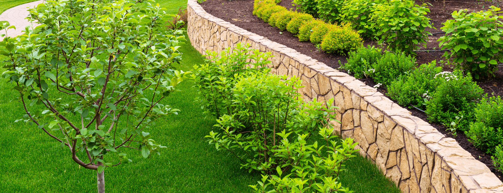 Image of a professionally designed and installed landscaping were they retaining wall several trees in small shrubs and green grass