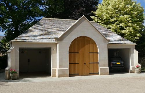 Stone garage with tiled roof, wooden arched door in center, opening on left, and car parked on right.